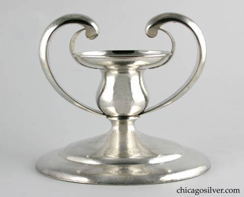 Kalo candlestick, massive, squat, with wide round base, flat bottom, very large central bobeche with oversize upturned rim and wire applied to rim.  Two large arms curve upward from the neck then over and down to join two wide curved strap handles that attach to the underside of the rim in a sort of heart-shape.  Engraved ' "Eleanor" / In Memory of / E. Louise Goodhue / 1933' on top of base.  Nice hammering, and heavy.  7-1/2" W at base (7-1/8" W across handles) and 6-1/2" H.  Marked:  STERLING / HAND WROUGHT / AT / THE KALO SHOP / C50 / FILLED
