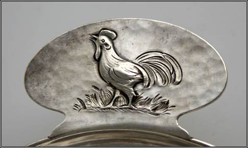 Kalo porringer, round, flat bottom form with tab handle and applied wire rim.  Handle has chased and repouss decoration of rooster standing on grass.  Detail shown.