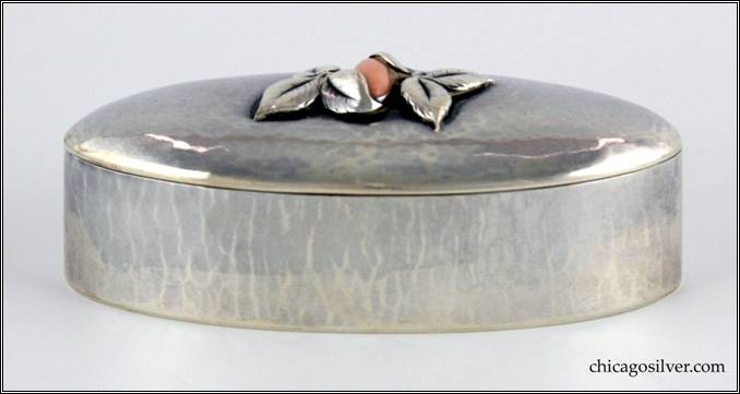 Kalo box, oval, covered, slender, with domed cover which has a bezel-set cabochon of pink coral flanked by two clusters of leaves.  Hammered surfaces with elongated hammer marks on the sides.  3-1/2" W and 1-3/8" H.  Marked:  STERLING / KALO