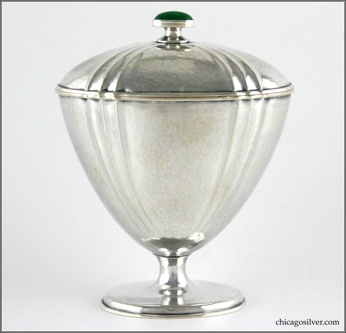 Kalo urn, covered, small, oval, with jade finial.  Wide mouth tapering to small stem on oval stepped foot, with pattern of three outward-pointing flutes repeated four times around the sides and in a matching arrangement on the removable cover.  Applied wire around top of urn and bottom of cover.  Cover has small square key on bottom of rim that fits into notch in rim of urn. Oval green jade bezel-set finial on cover.  Nicely hammered surfaces.  3-13/16" L and 2-1/2" W and 4-13/16" H.  Marked:  456 M / STERLING / HAND WROUGHT / AT / THE KALO SHOPS / CHICAGO / AND / NEW YORK